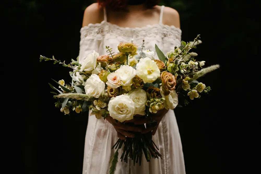 Novia de pie con un ramo de rosas blancas y amarillo pálido, sobre fondo oscuro.