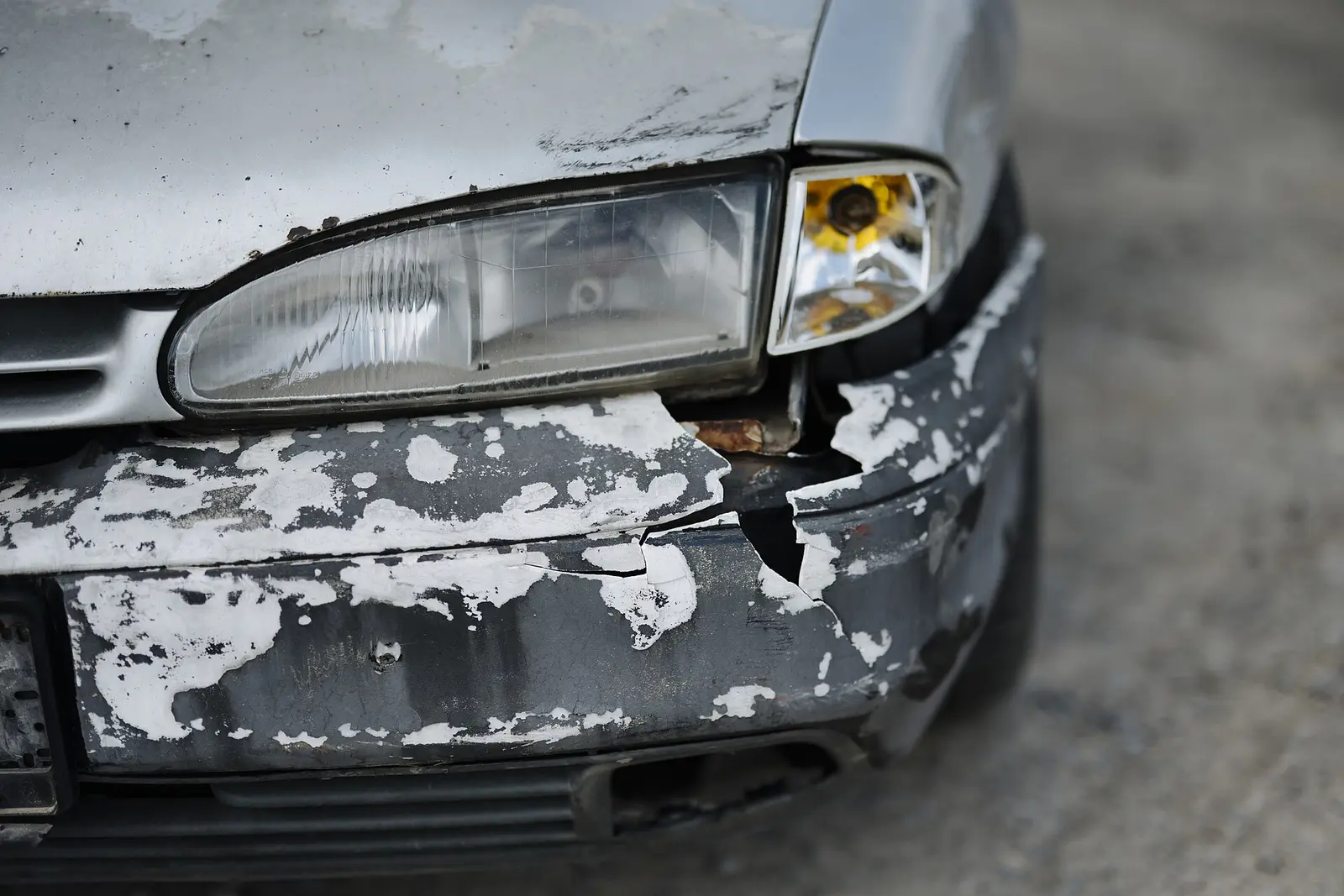 Frontal de un coche gris golpeado, con el paragolpes roto, la pintura levantada y el faro descolgado
