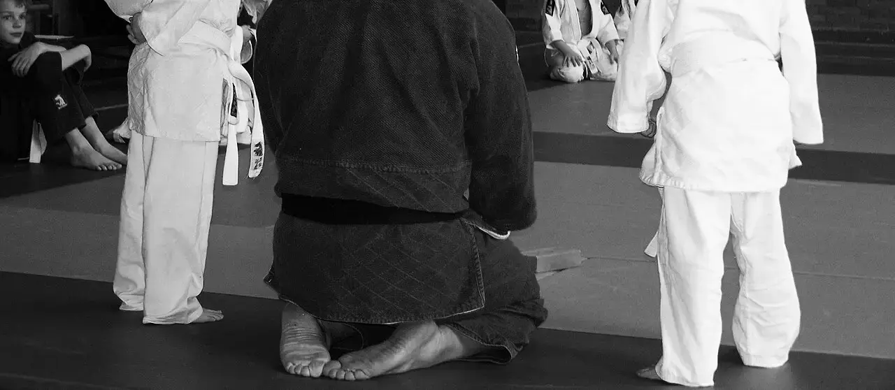Entrenador con kimono azul de rodillas en el tatami frente a dos niñas con kimono blanco, vistos de cintura para abajo