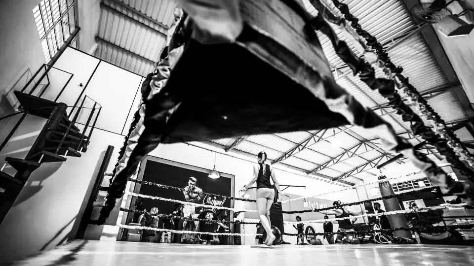 Sala de boxeo con un ring al fondo y una boxeadora calentando junto a las cuerdas