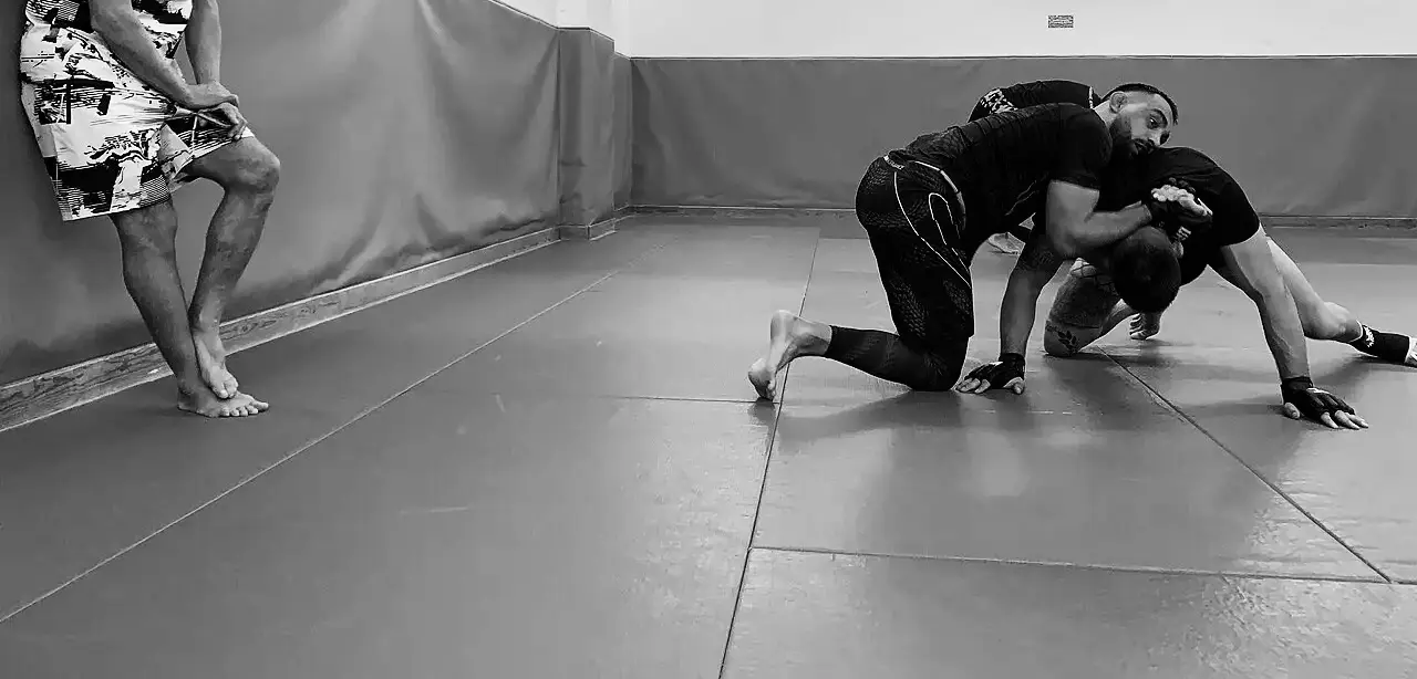 Dos personas entrenando jiu-jitsu sobre el tatami azul sobre el tatami de la sala, con el entrenador de pie a un lado