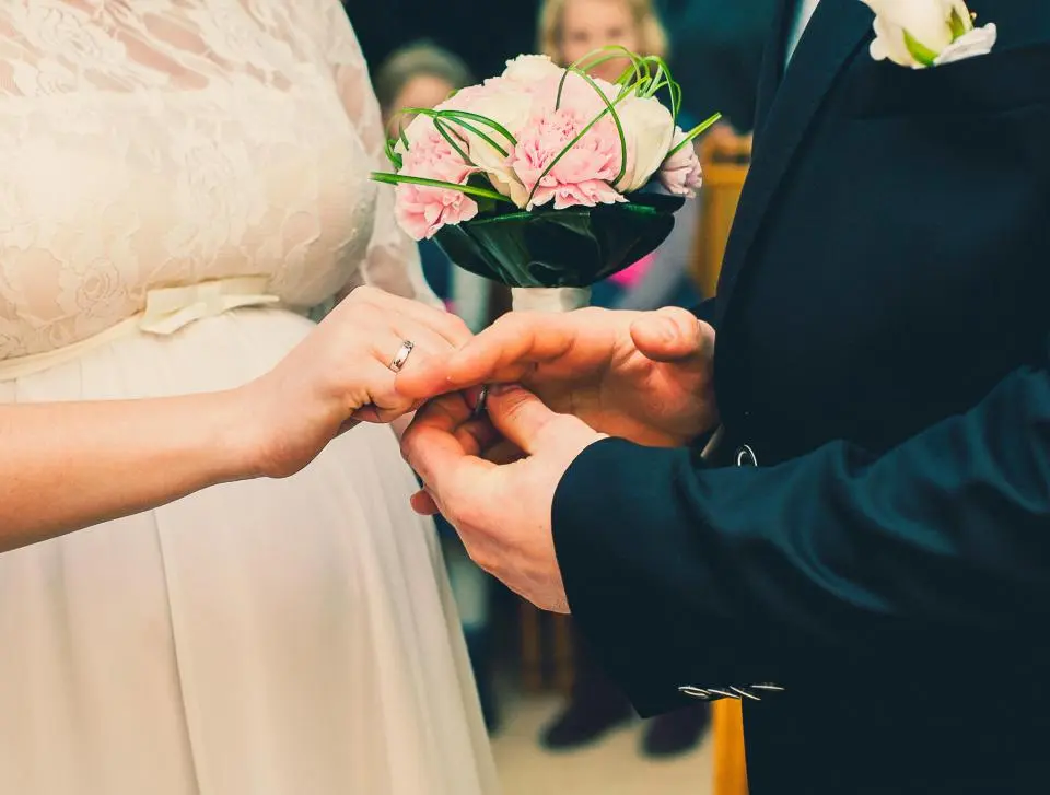El novio pone la alianza a la novia, que sujeta un ramo rosa