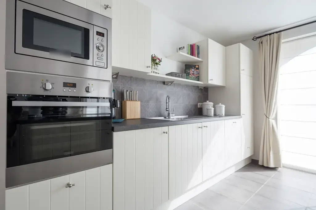 Cocina lineal blanca con horno en columna, frente gris y ventana con cortina