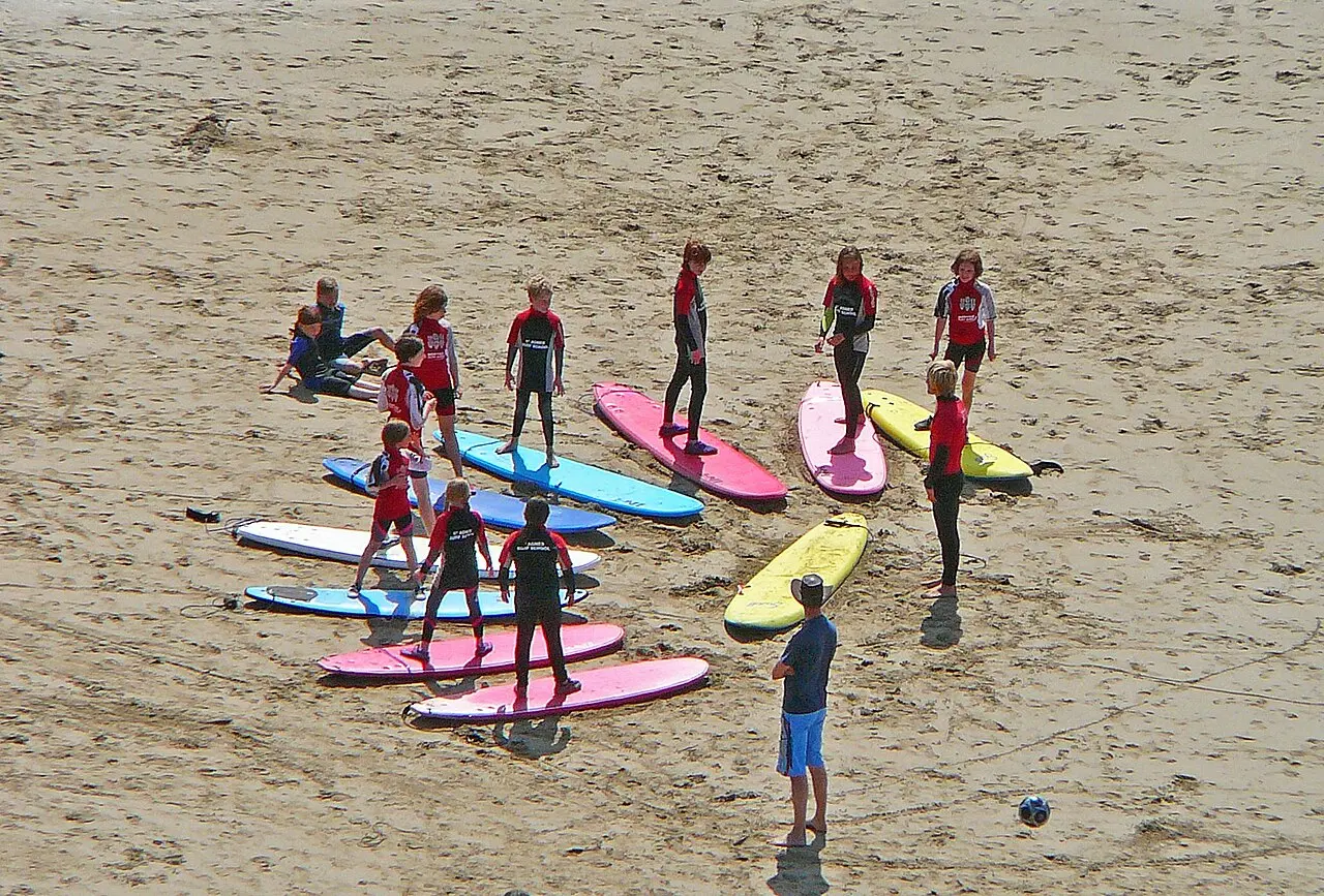 Un grupo de niños con neopreno practica de pie sobre tablas de colores en la arena, con el monitor delante.