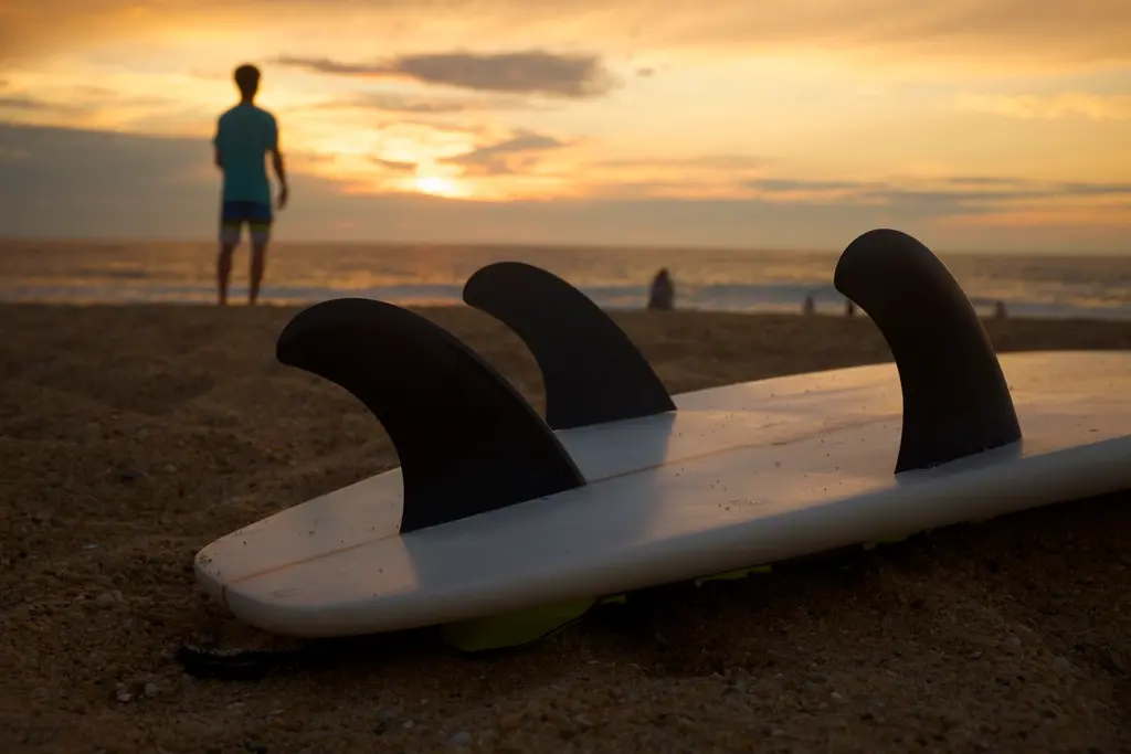 Una tabla de surf en la arena al atardecer, con las quillas hacia arriba y un surfista mirando el mar.