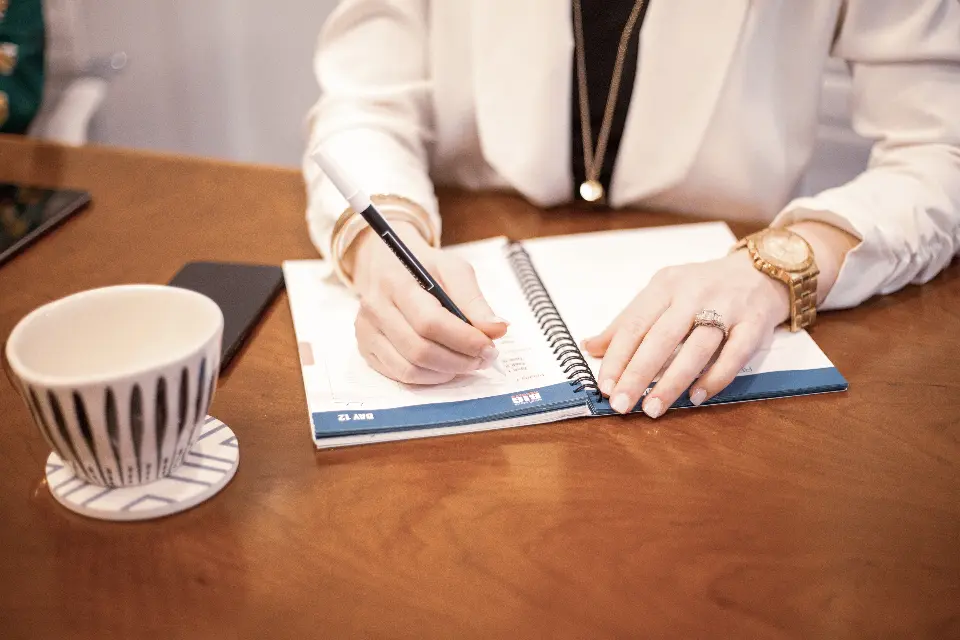 Manos escribiendo en una agenda sobre una mesa de madera, junto a una taza.