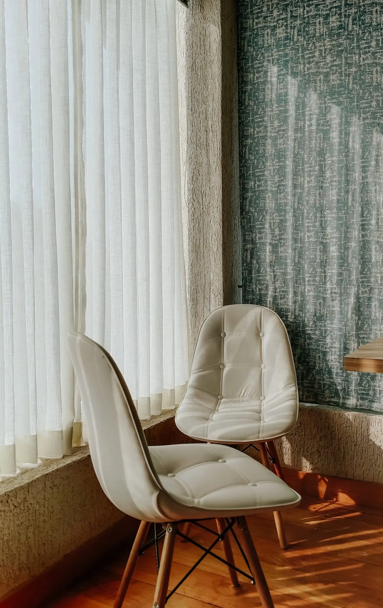 Dos sillas tapizadas en blanco junto a una ventana con cortinas finas y luz de tarde sobre el suelo de madera.