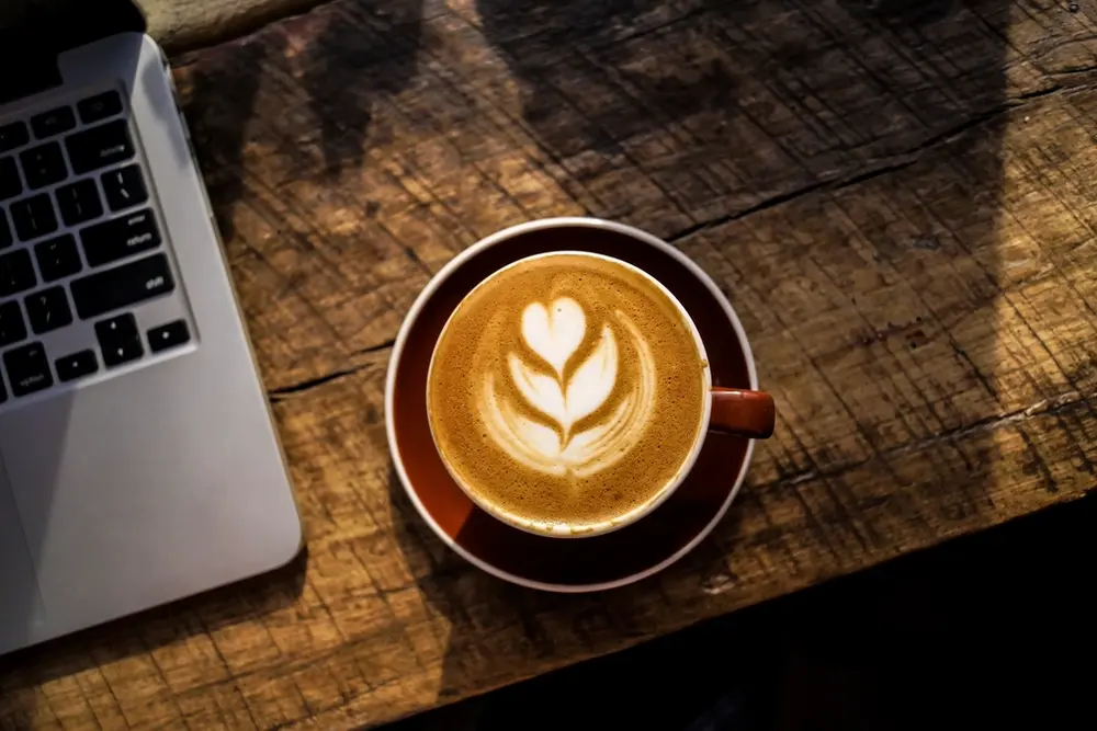 Café con dibujo de hoja junto a un portátil sobre la mesa de madera