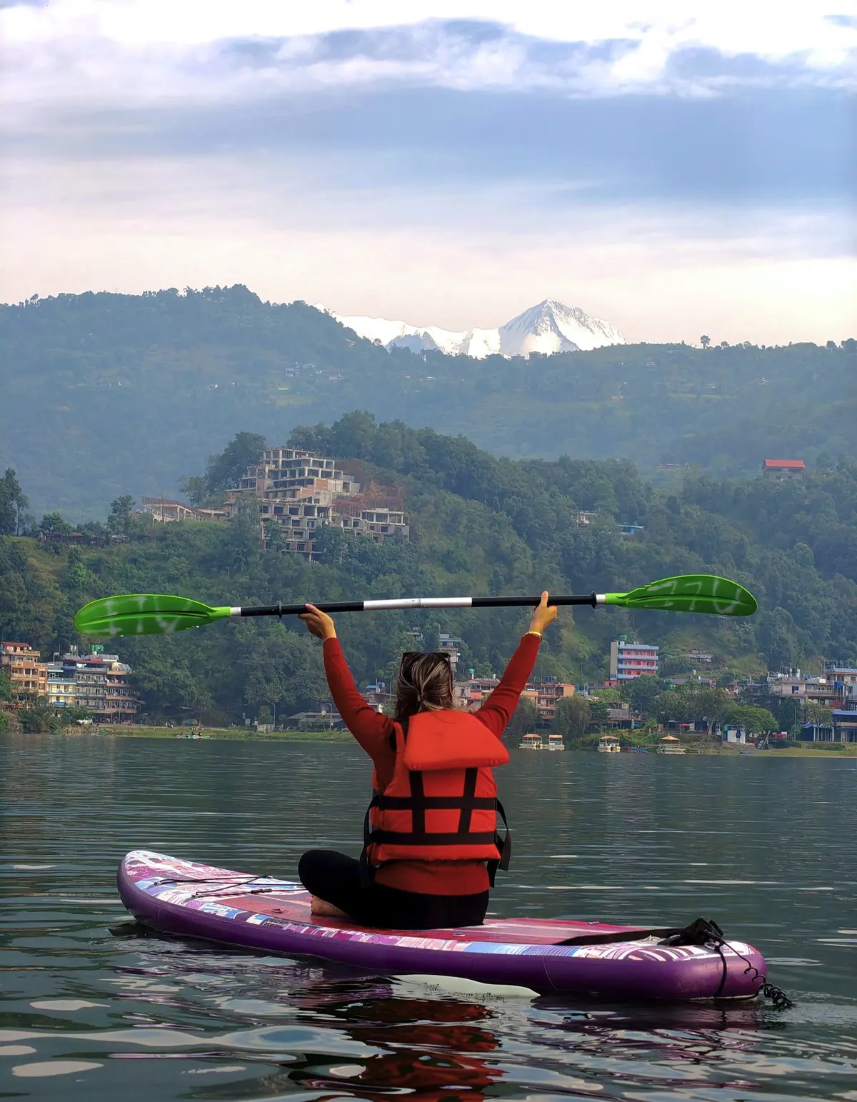 Una chica sentada en una tabla de paddle surf levanta la pala, en un lago rodeado de monte.