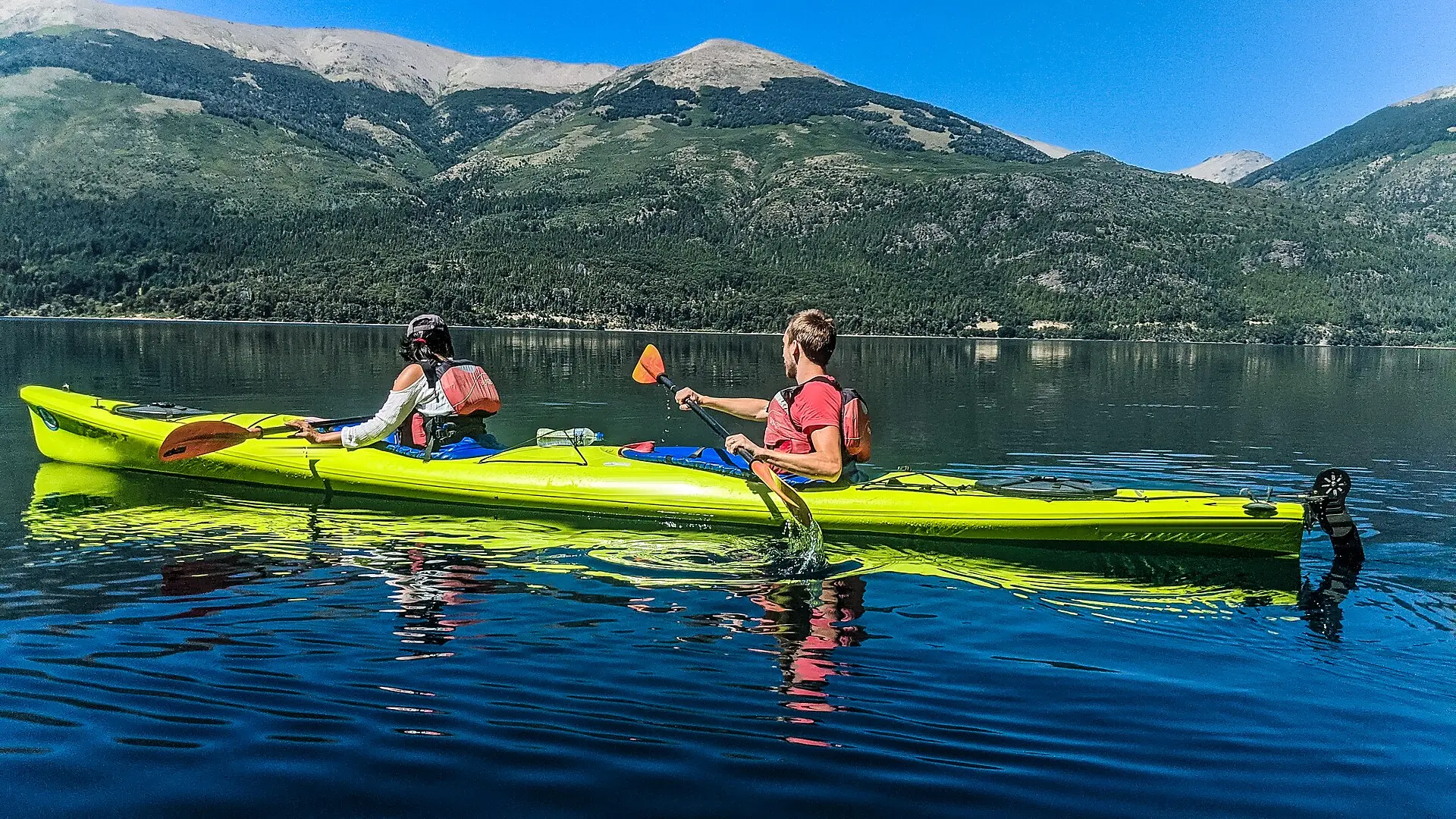 Dos personas reman en un kayak doble amarillo por un embalse en calma, con montañas verdes y cumbres peladas detrás.