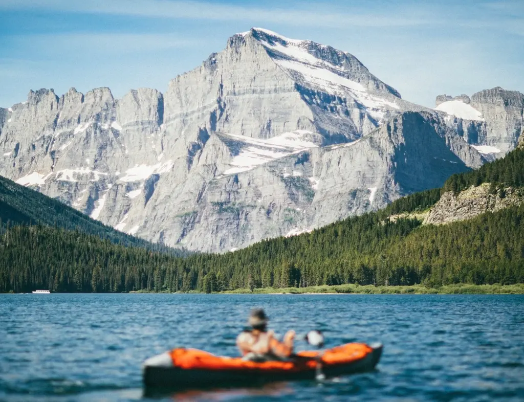 Una persona rema en un kayak naranja hacia unas montañas de roca gris, con el lago en calma.