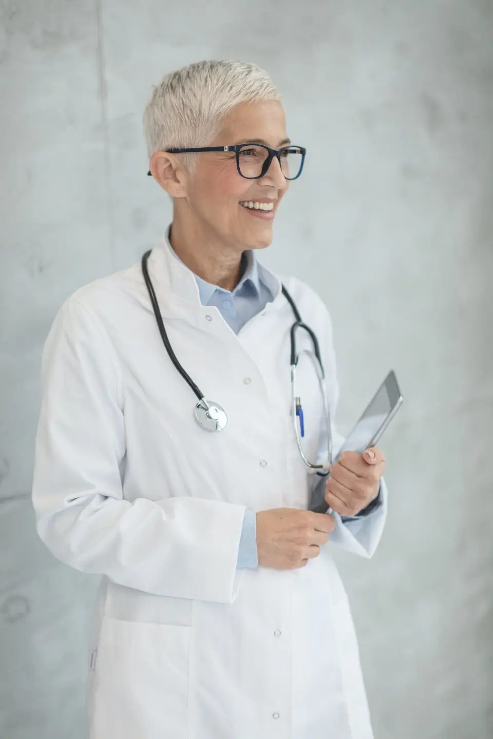 Una sanitaria con bata blanca y gafas sonríe con una carpeta en la mano.