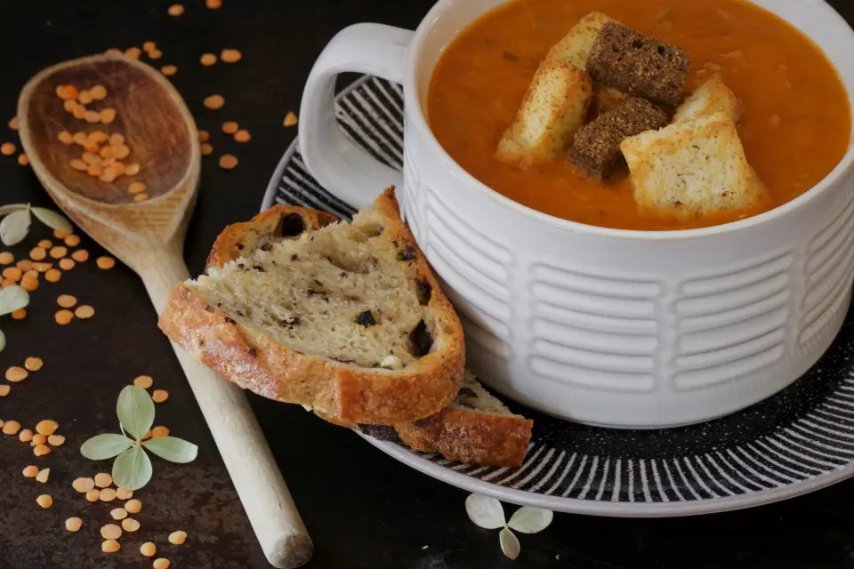 Una taza de crema de lentejas con picatostes, pan y una cuchara de madera.