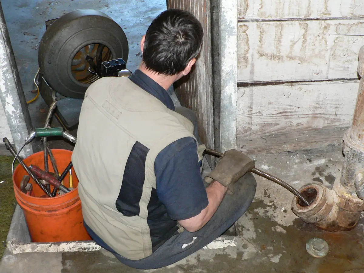 Fontanero de espaldas, sentado en el suelo, desatascando una bajante con la máquina de muelle.