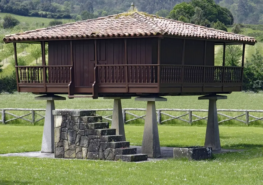 Hórreo de madera oscura con tejado de teja sobre cuatro pilares de piedra, con su escalera de piedra, en medio de un prado