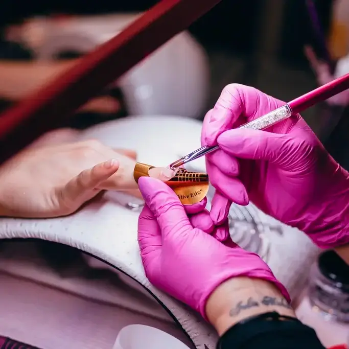Manos con guantes rosas pintando con pincel las uñas de una clienta, junto a la lámpara